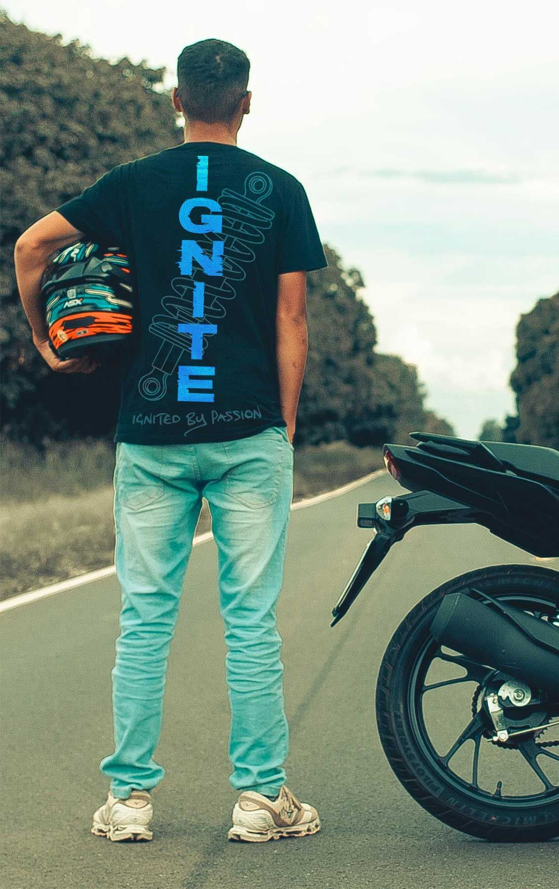 Man standing on a road beside a motorcycle, wearing a black IGNITE shirt and light jeans.