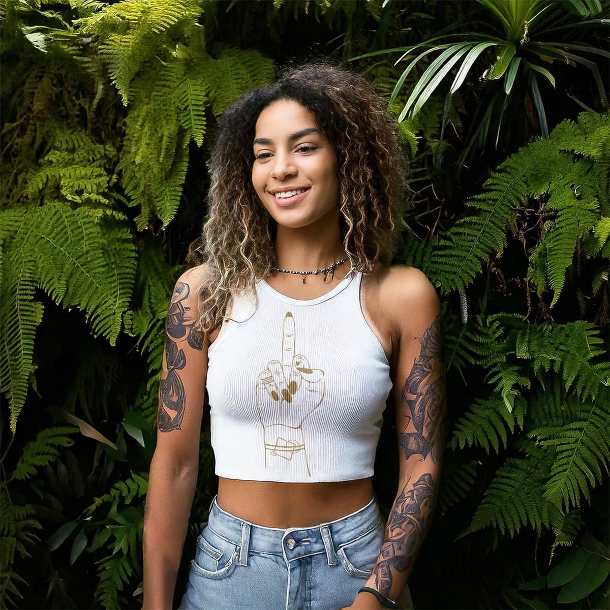 Smiling woman with tattoos in a white crop top and jeans, standing before lush green foliage