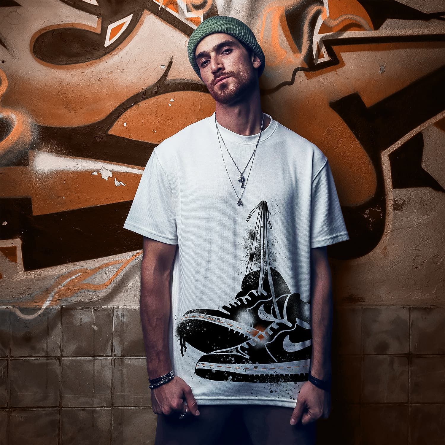 Man standing against a graffiti wall, wearing a white graphic T-shirt and green knit beanie.
