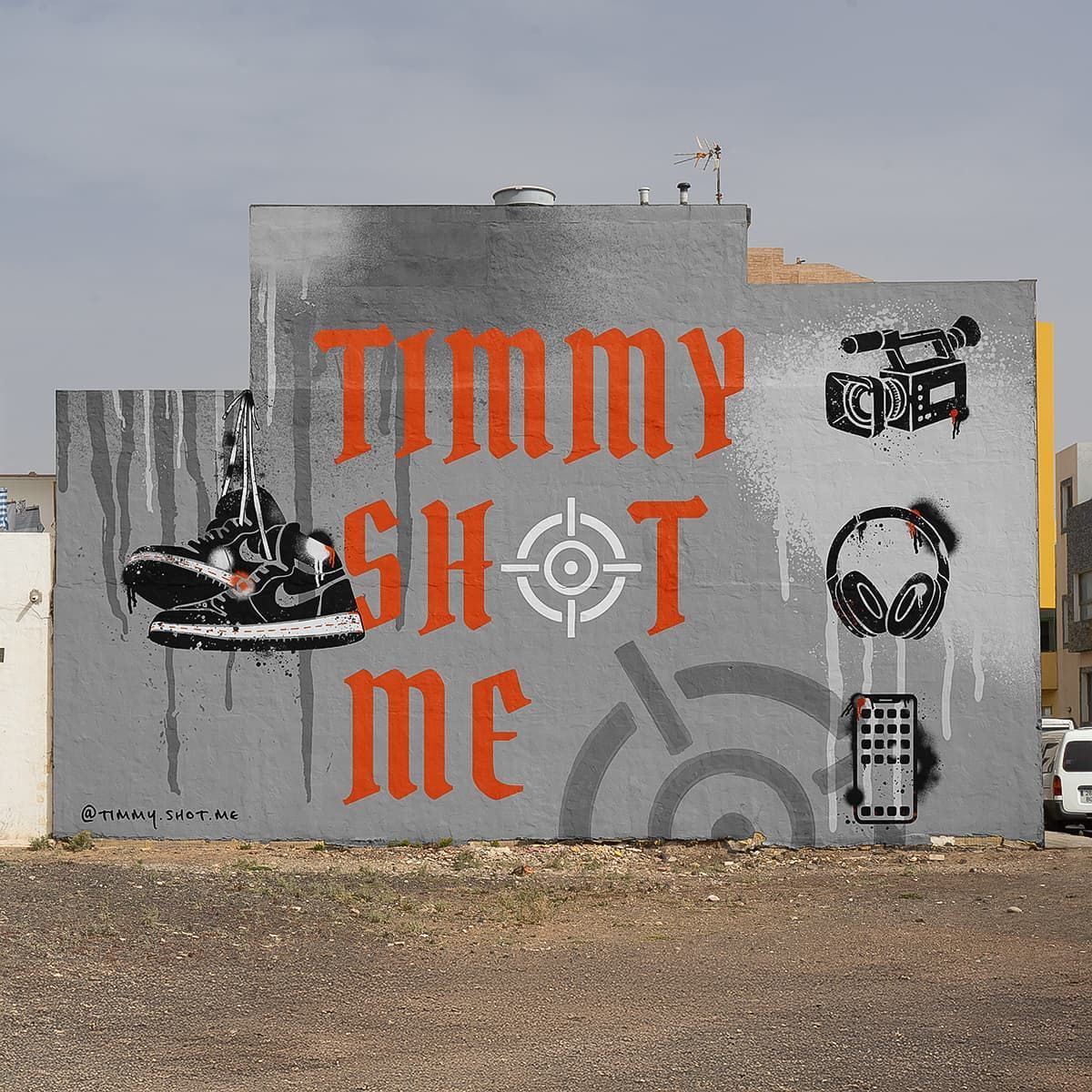 Gray wall mural with orange “TIMMY SHOT ME” text and black graffiti icons in a gravel lot