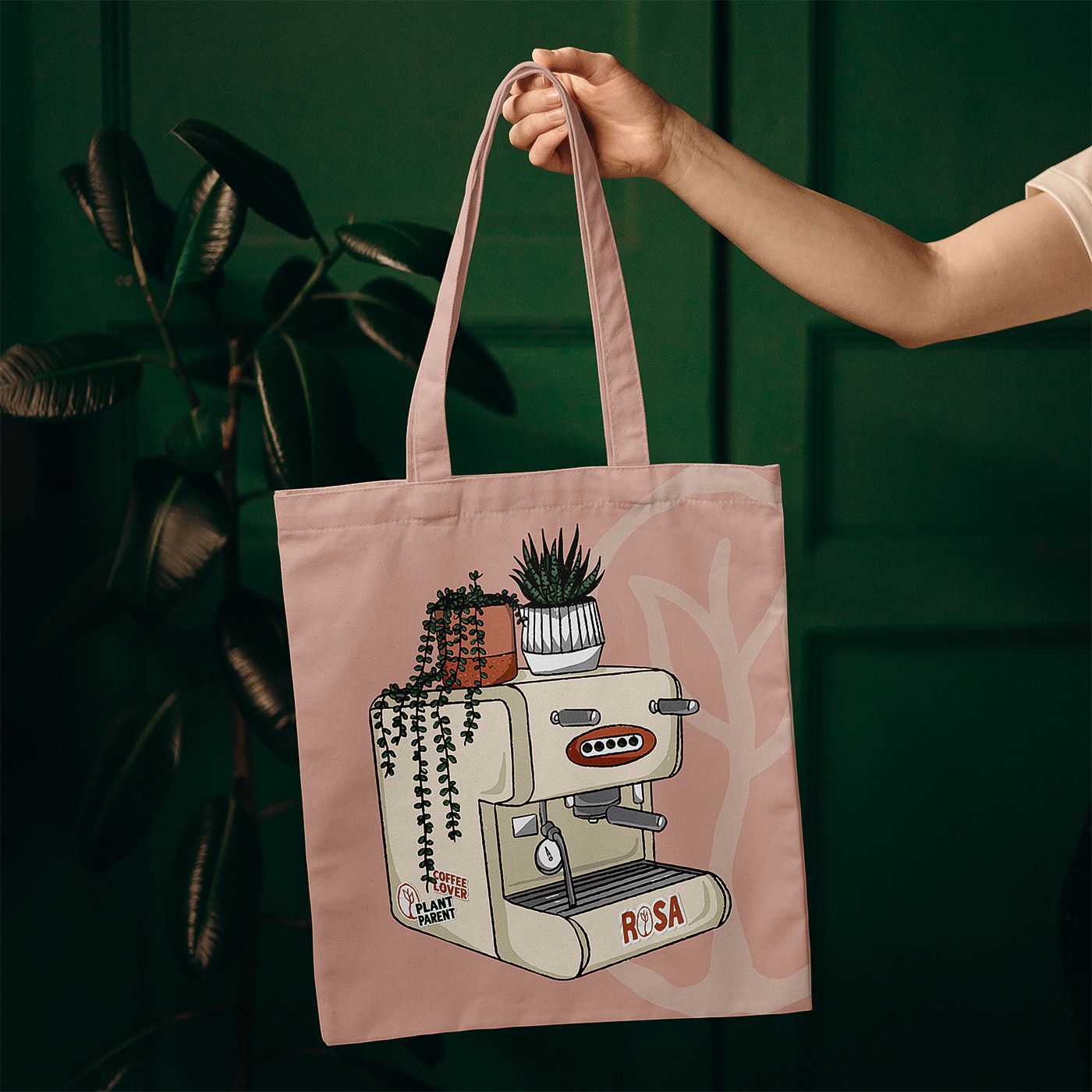 Hand holding a pink tote bag with a cat illustration against a dark green background