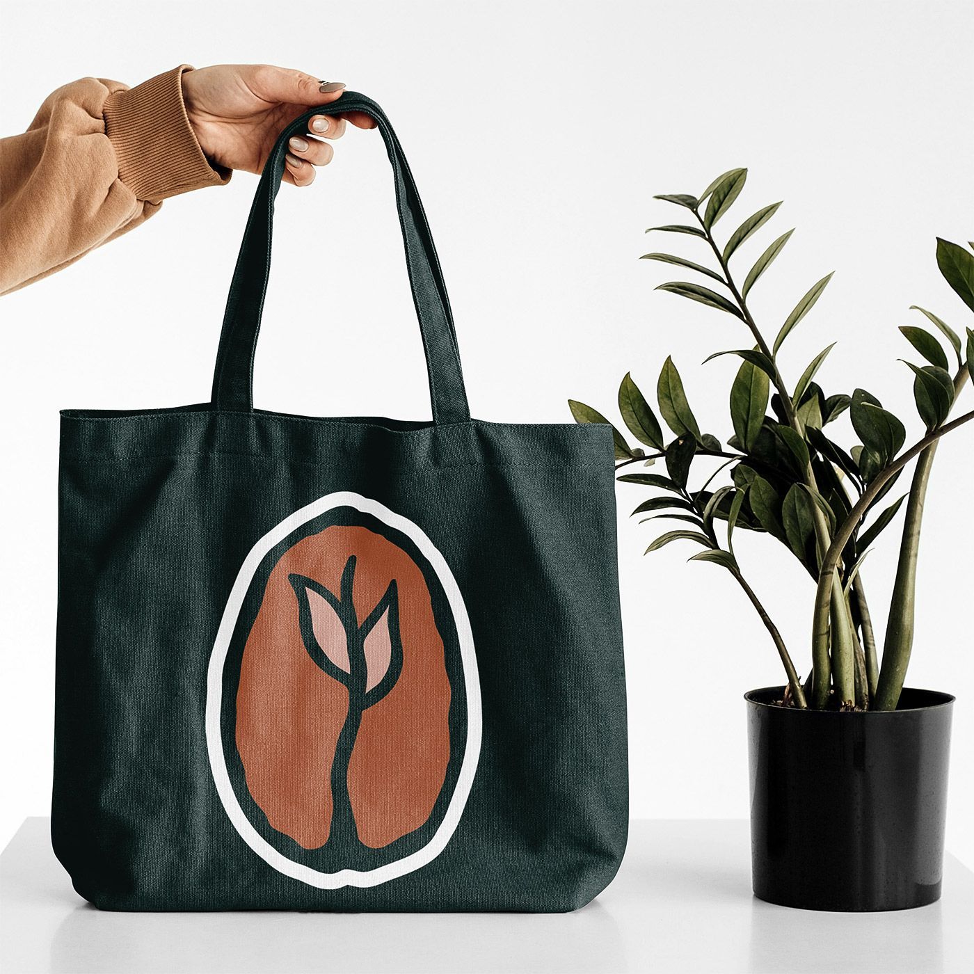 Black tote bag with orange leaf logo held beside a potted plant on a white background