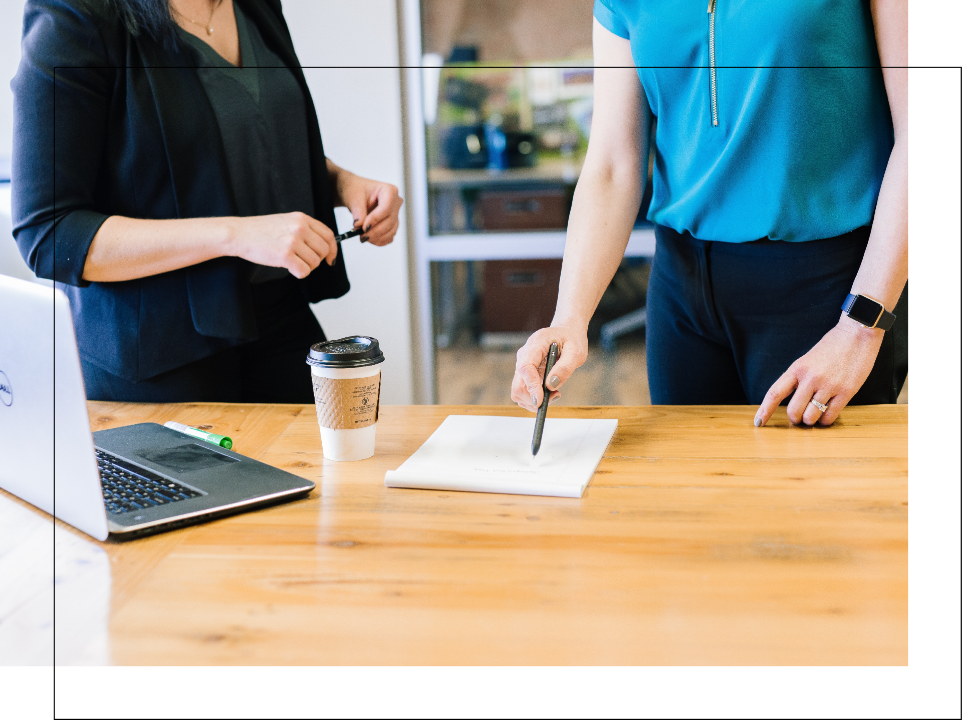 Two women are standing at a table with a laptop and a cup of coffee.