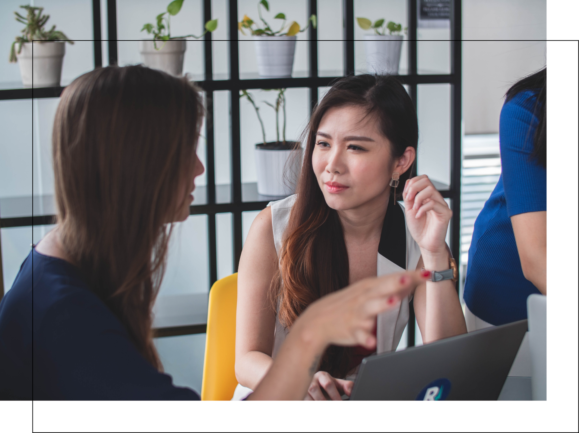 Two women are sitting at a table with a laptop and talking to each other.