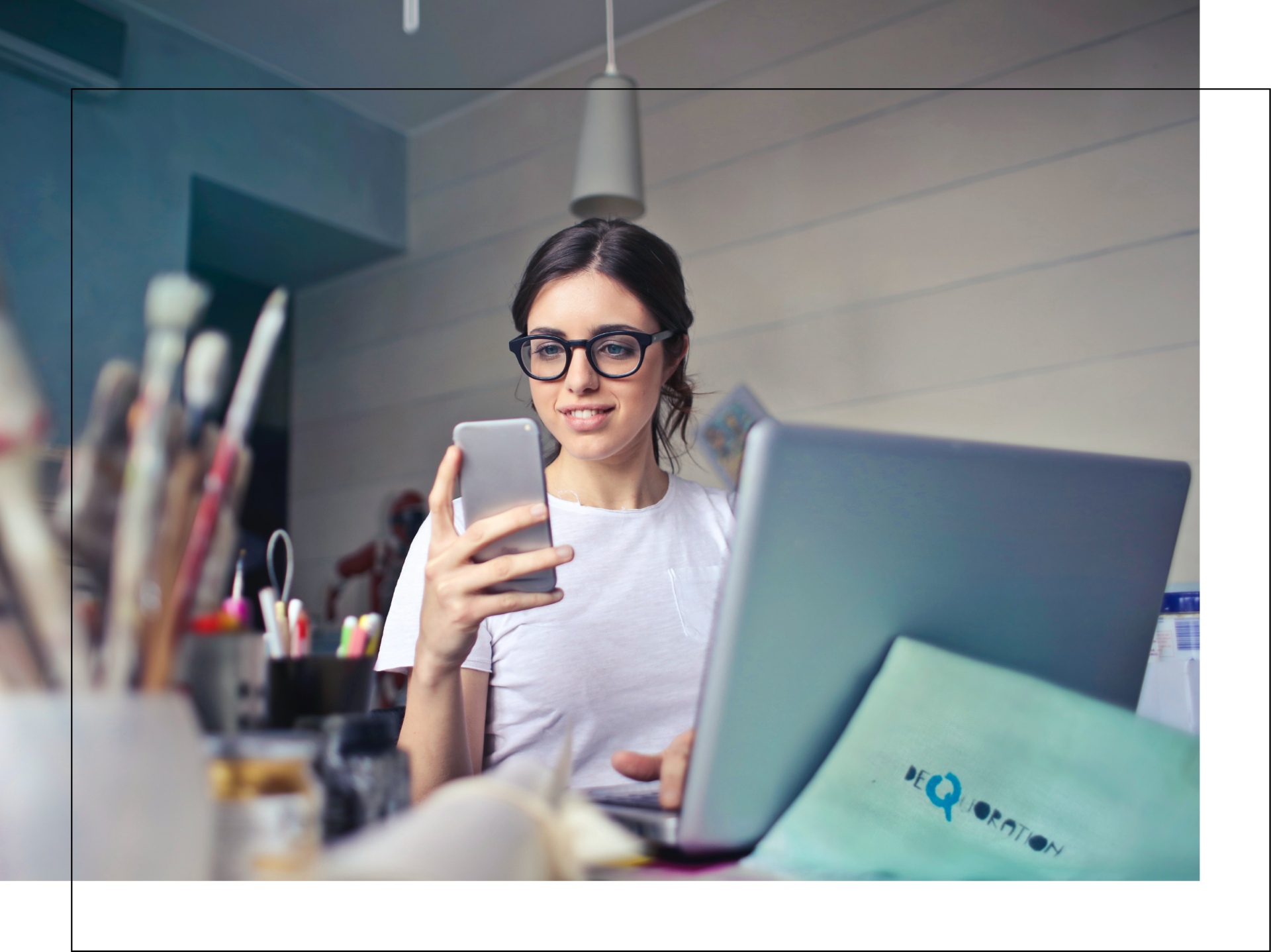 A woman is sitting at a desk using a laptop and a cell phone.