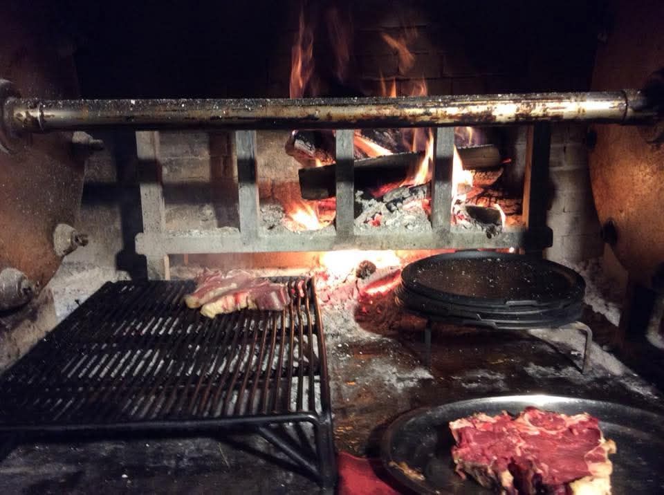 Camino in mattoni con braci ardenti, griglia metallica e una padella con della carne che cuoce all'interno.