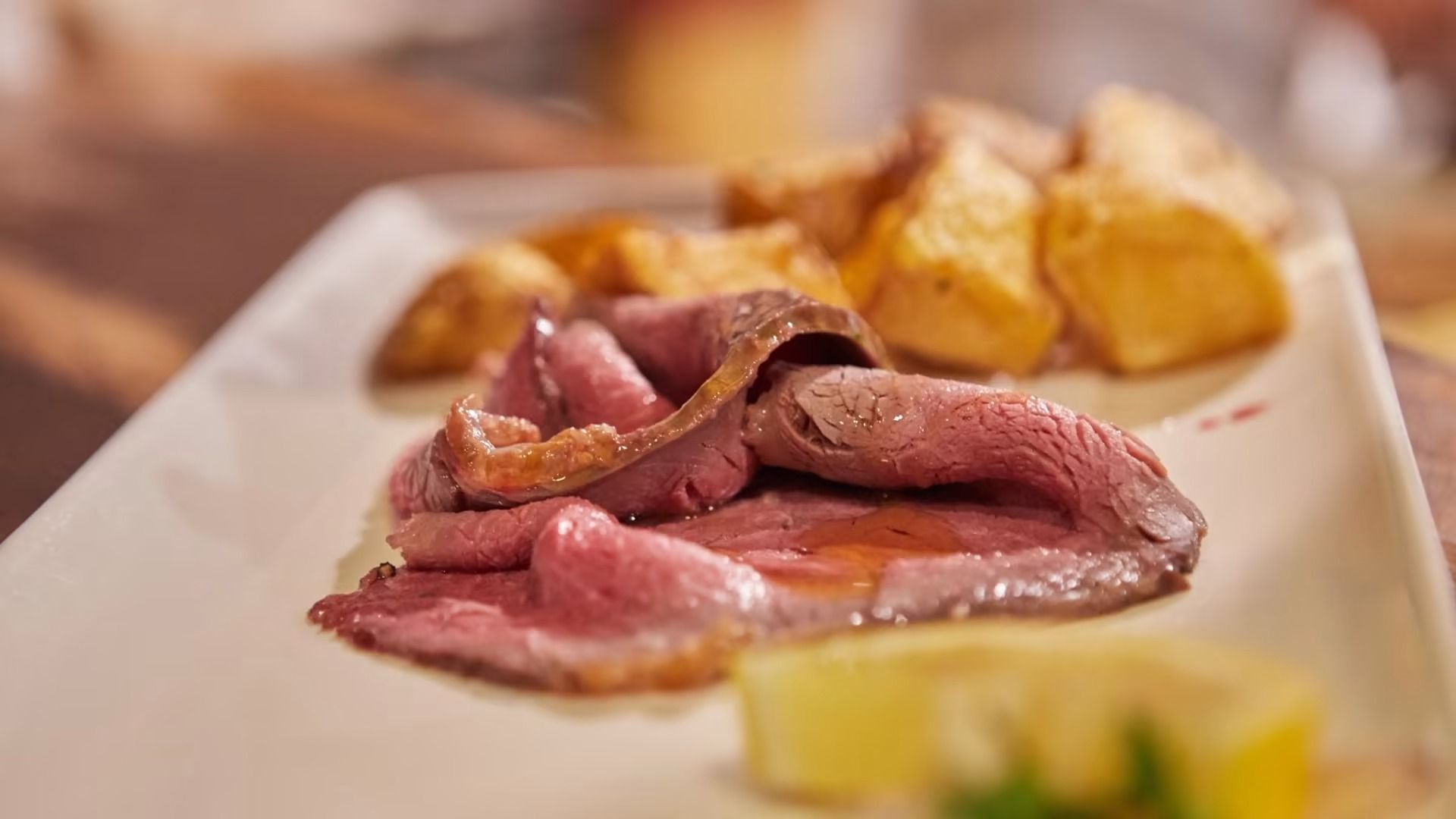 Roast beef affettato con patate arrosto e spicchio di limone su un piatto bianco.