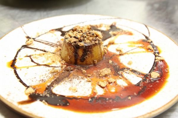 Dessert: budino circolare color caramello, ricoperto di briciole e salsa al cioccolato, servito su un piatto bianco.