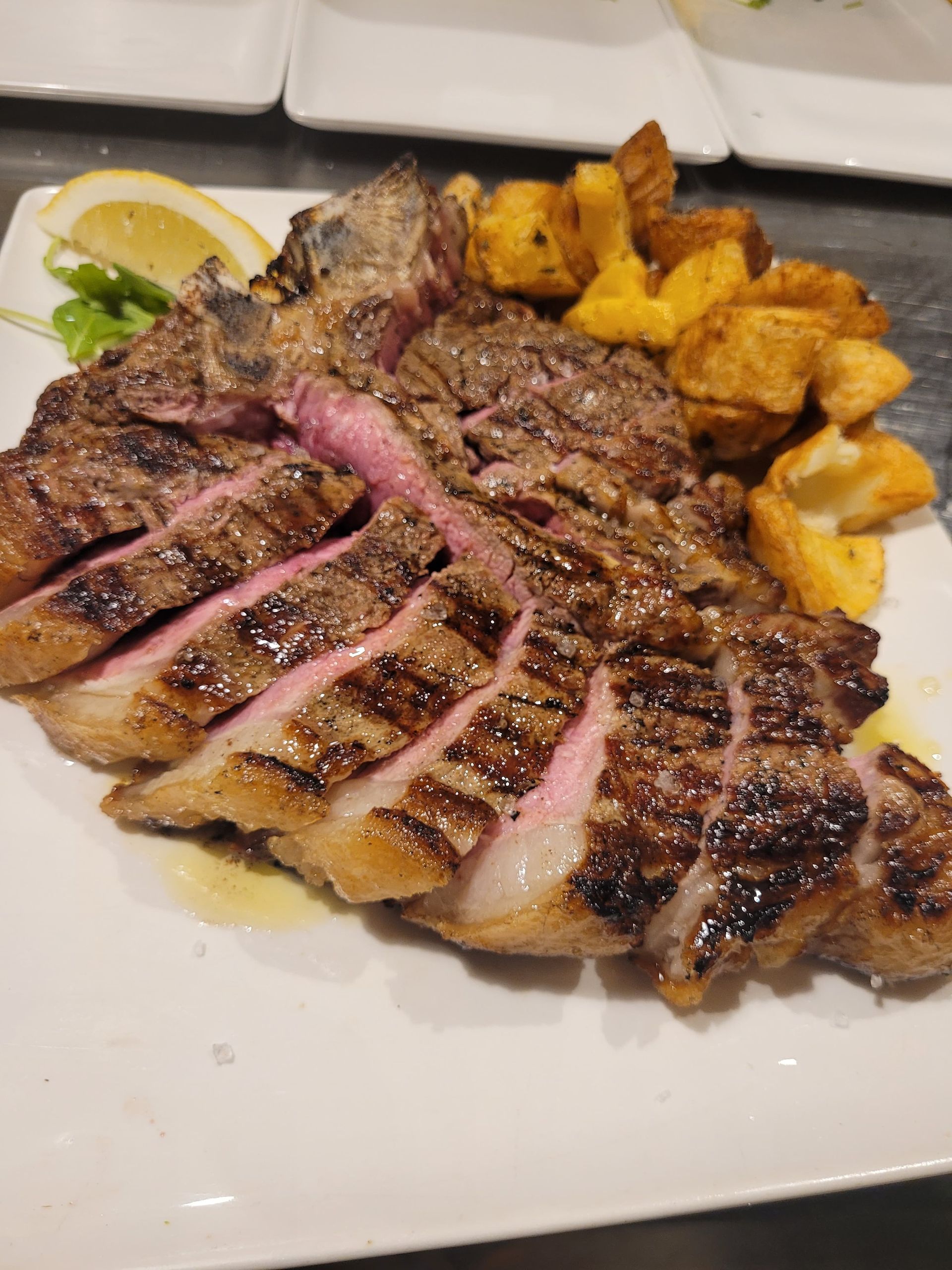Bistecca alla griglia tagliata a fette con patate e limone su un piatto bianco.
