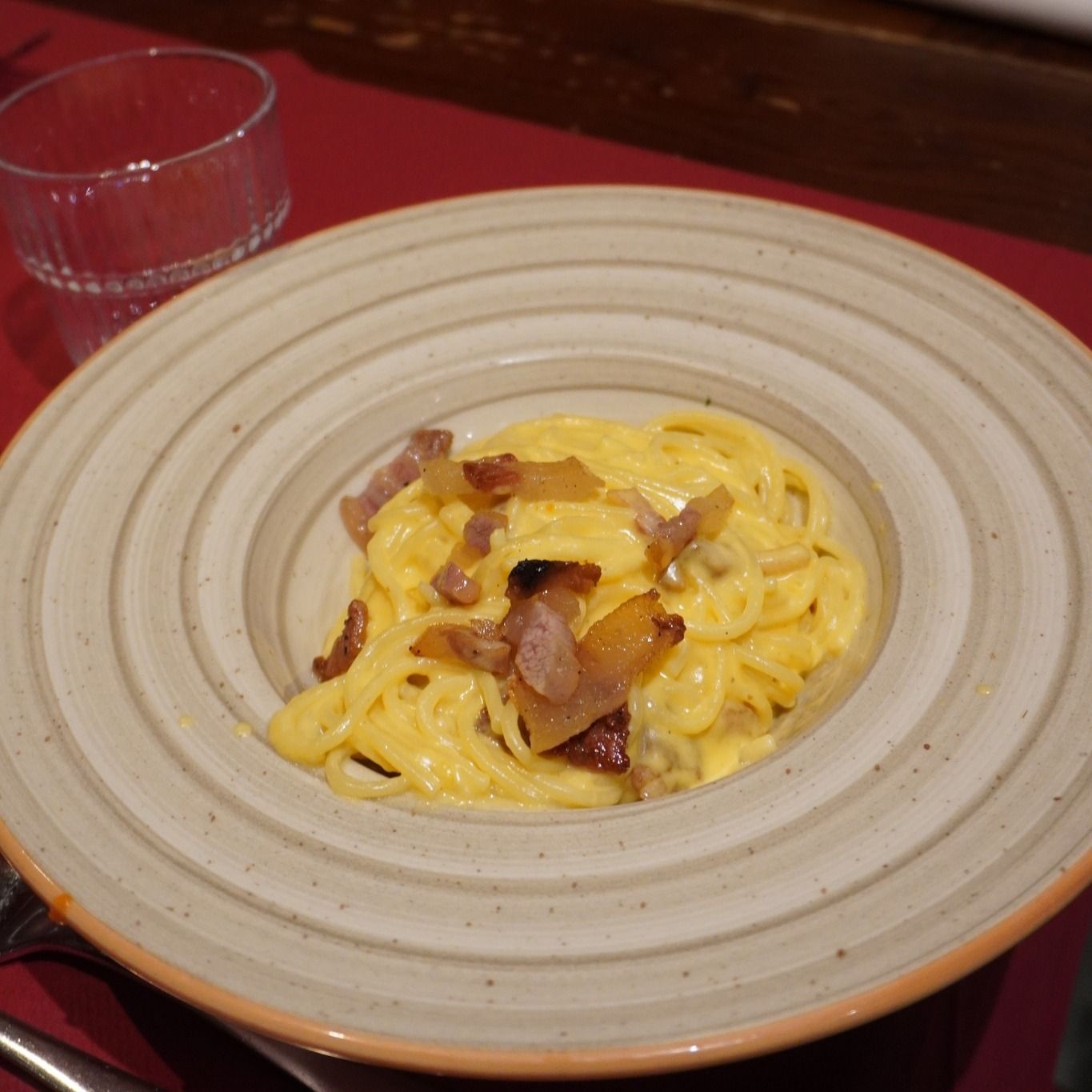 Pasta alla carbonara in una ciotola beige con pancetta su una tovaglia rossa.