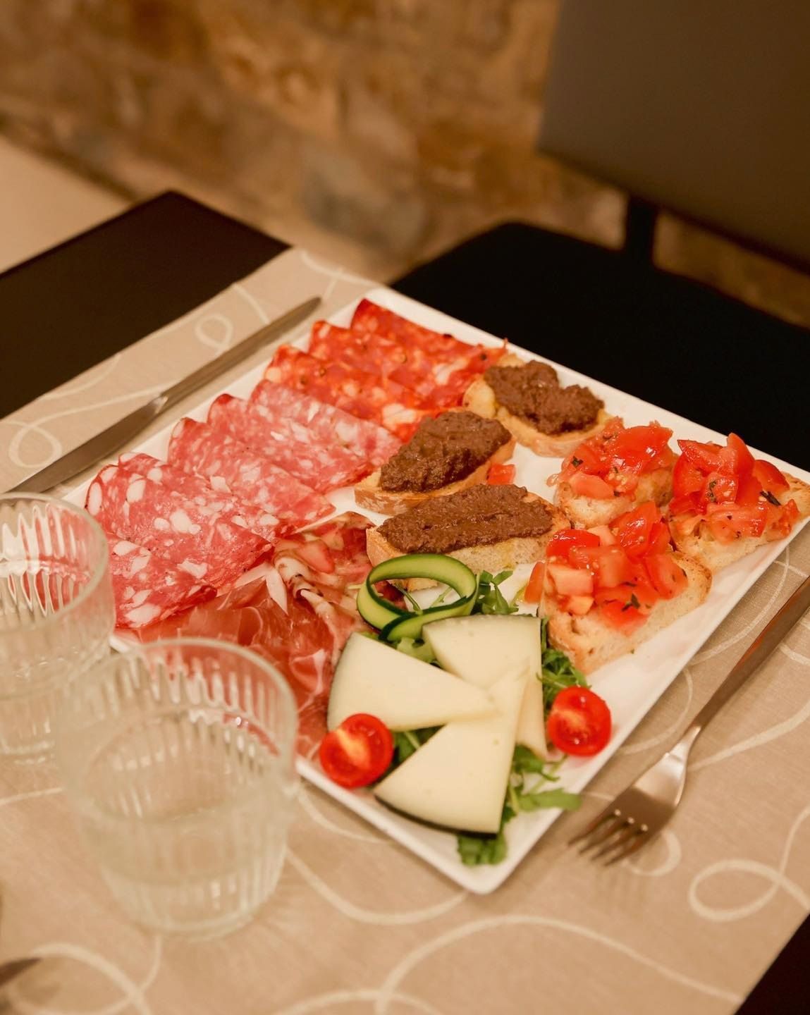 Piatto di salumi con affettati, formaggio, bruschetta e tapenade di olive, apparecchiato al tavolo.