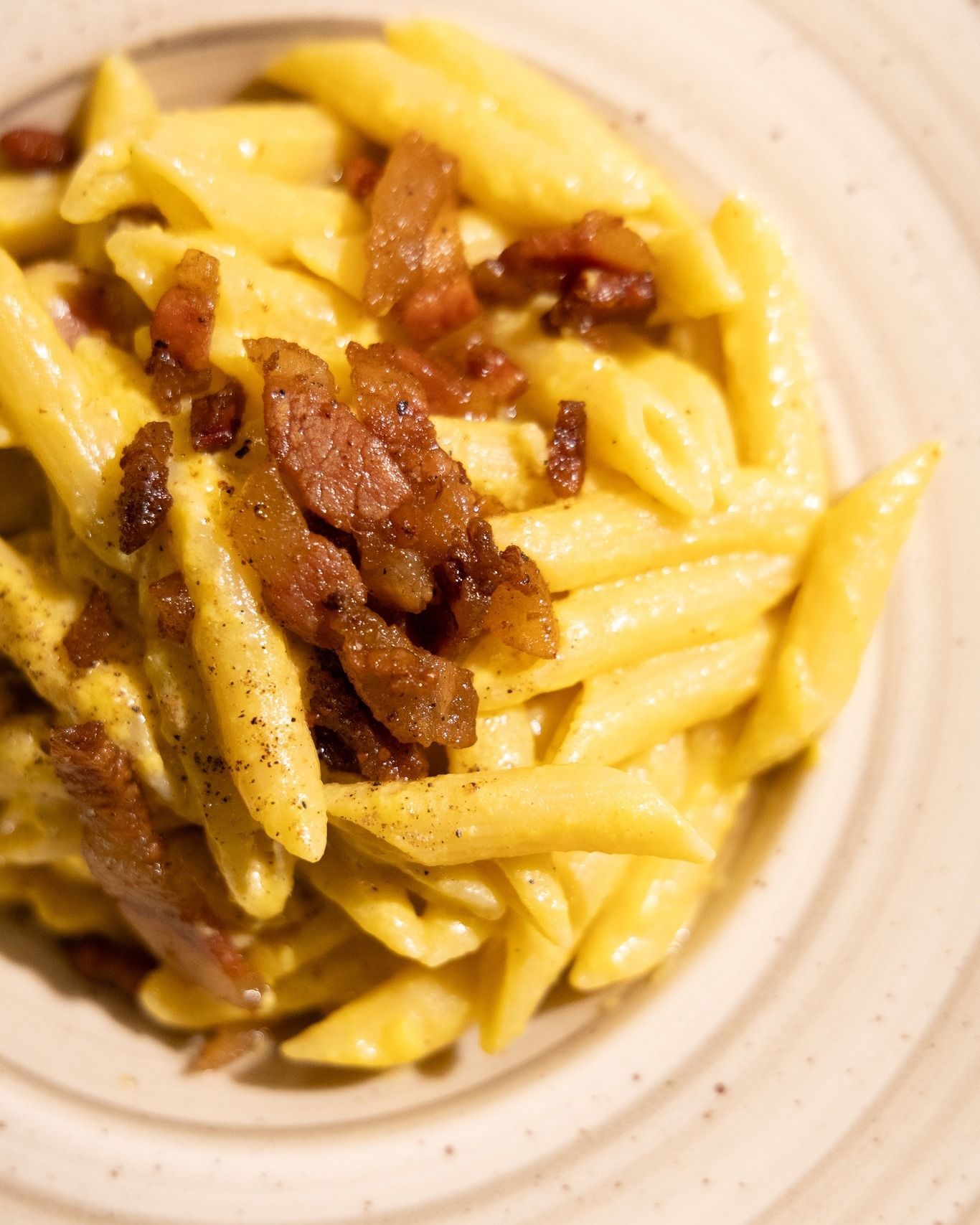 Penne condite con una cremosa salsa gialla, guarnite con pezzetti di pancetta croccante, in una ciotola bianca.