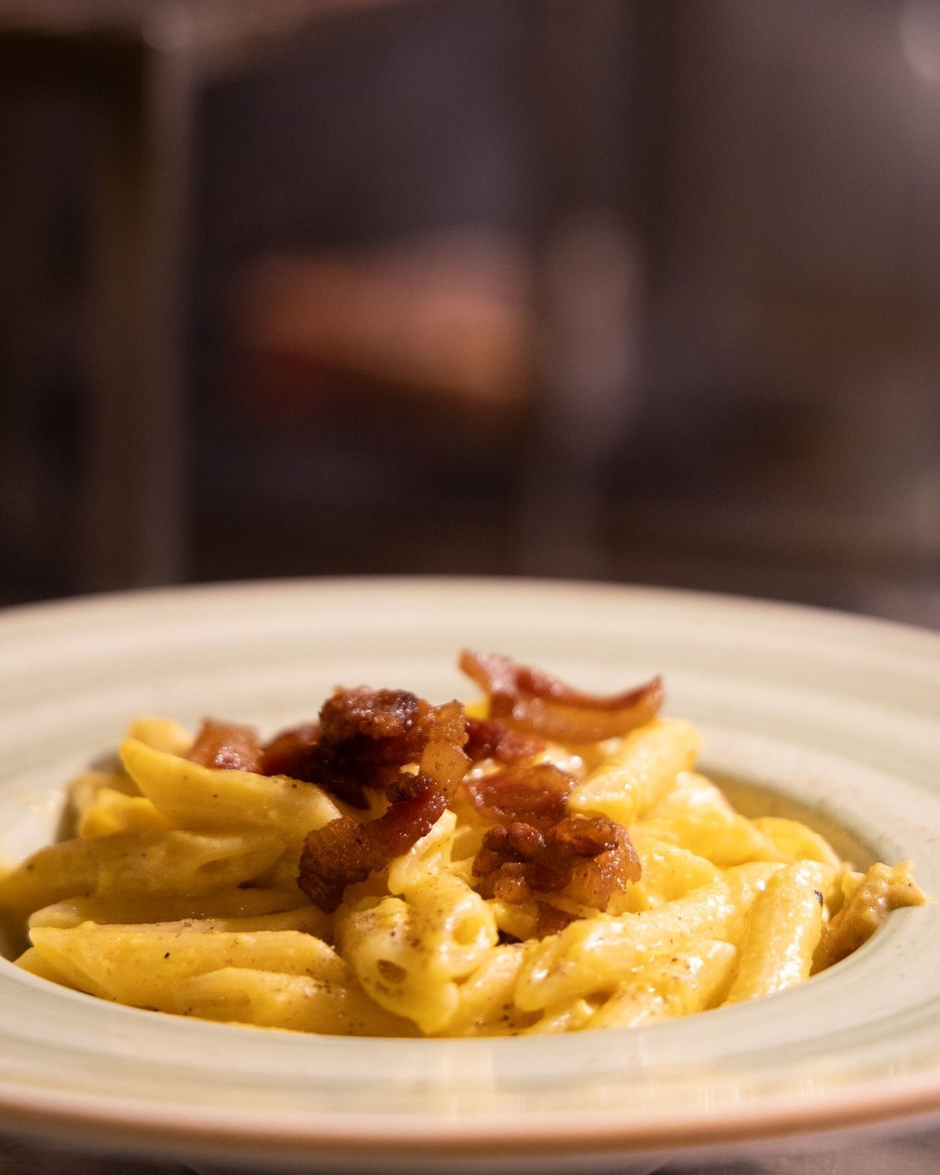 Penne condite con una cremosa salsa gialla, guarnite con pancetta croccante, in una ciotola con bordo verde.