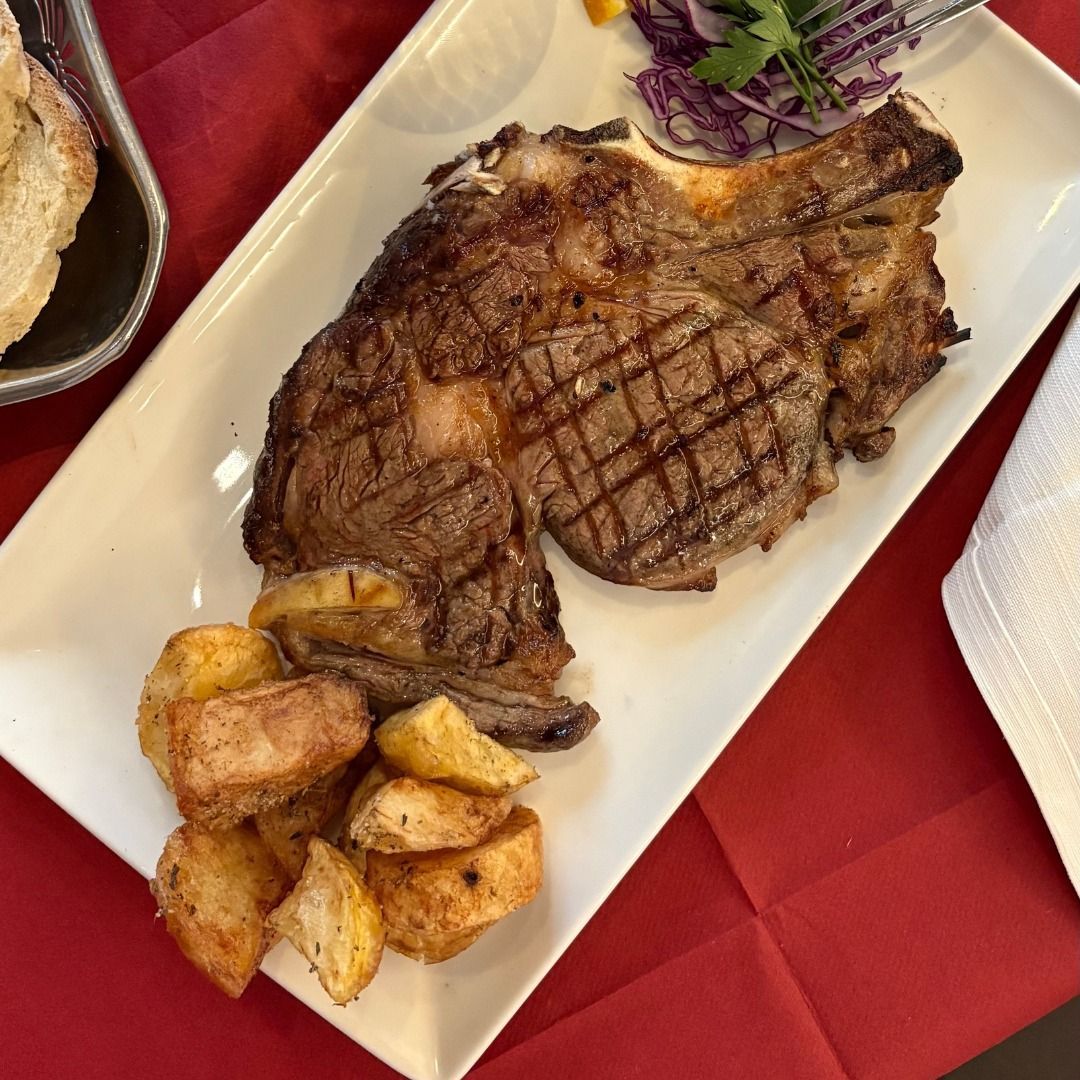 Bistecca alla griglia con patate e contorno su un piatto bianco, adagiato su una tovaglia rossa.