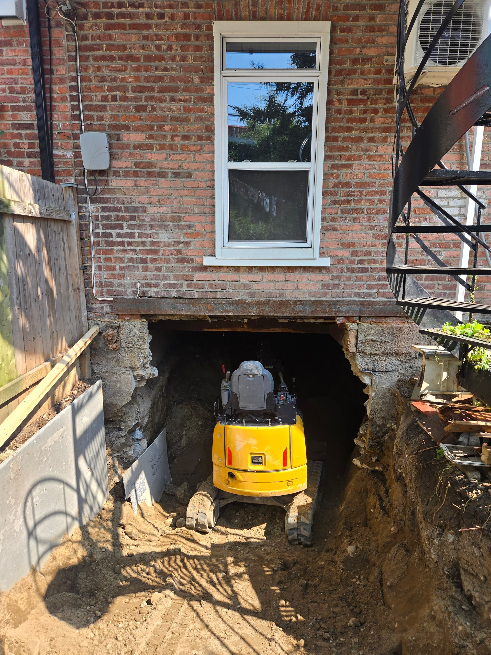 Mini-pelle jaune dans un sous-sol creusé sous une fenêtre de maison en briques