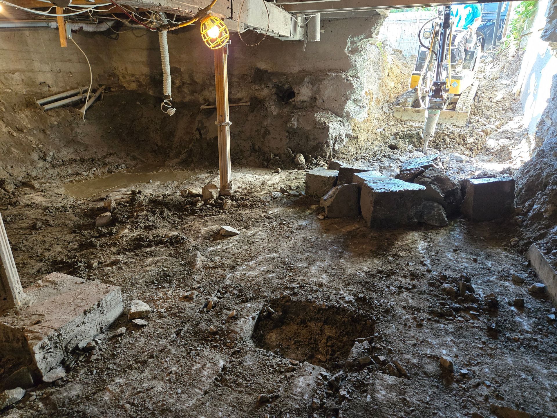 Fosse de fouille boueuse avec des pierres, un poteau central et une lampe suspendue sous un abri bas recouvert d'une bâche.