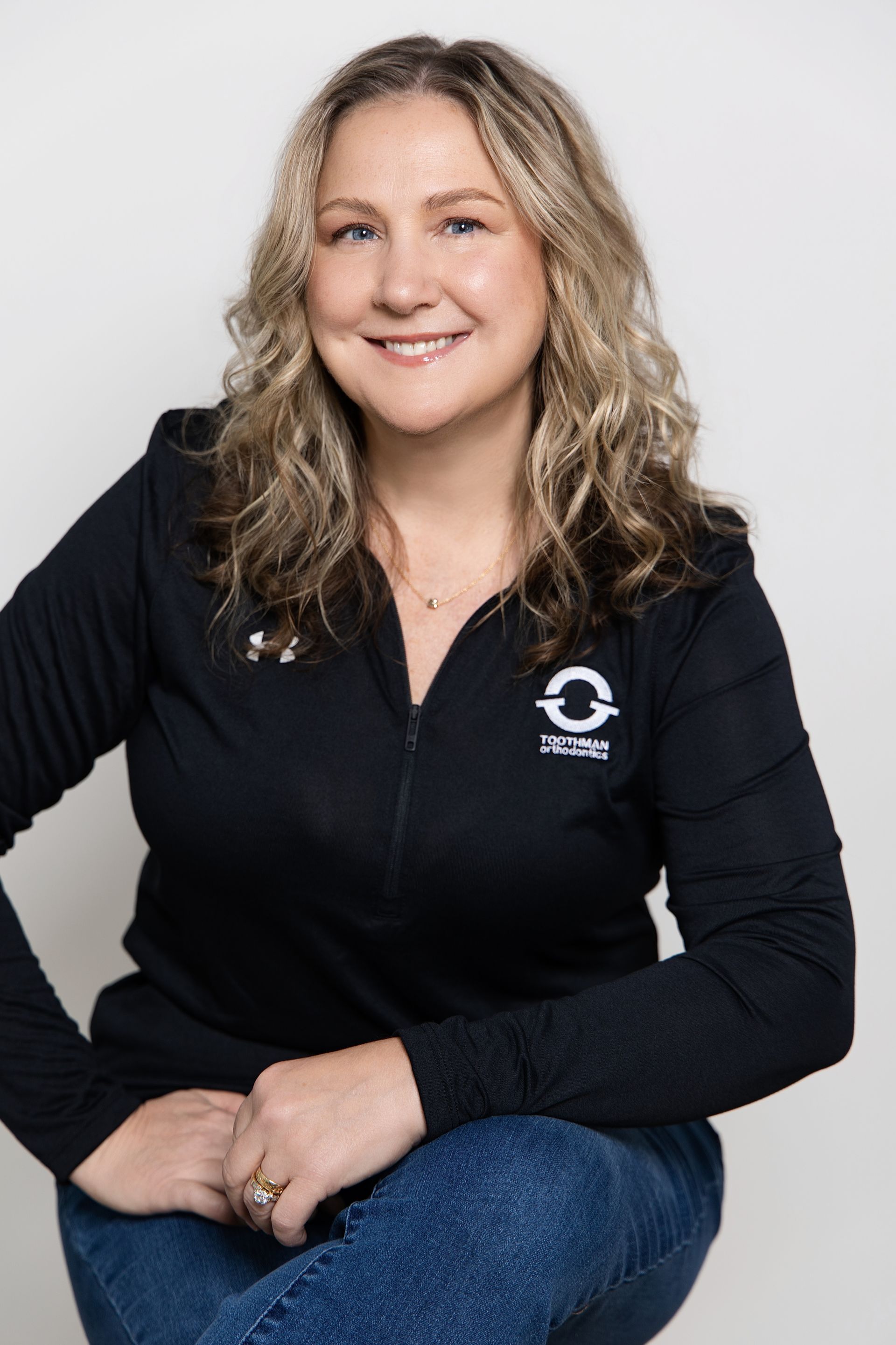 Smiling woman in a black zip-up top and jeans, seated against a light gray background.