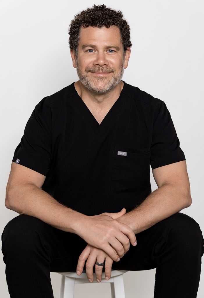 Man in a black shirt seated on a stool against a plain light background, smiling gently.