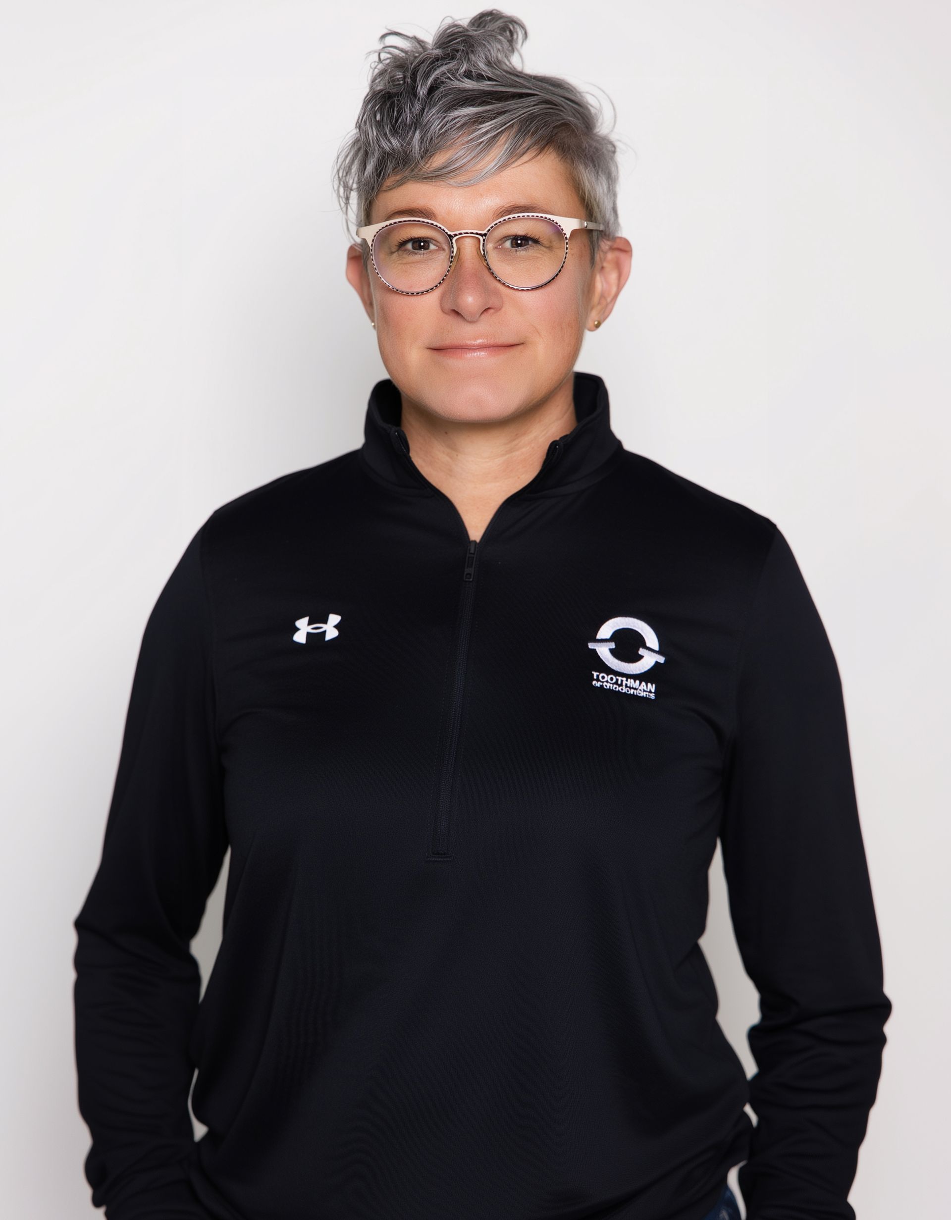 Portrait of a person with short gray hair and glasses in a black zip-up top on a plain background