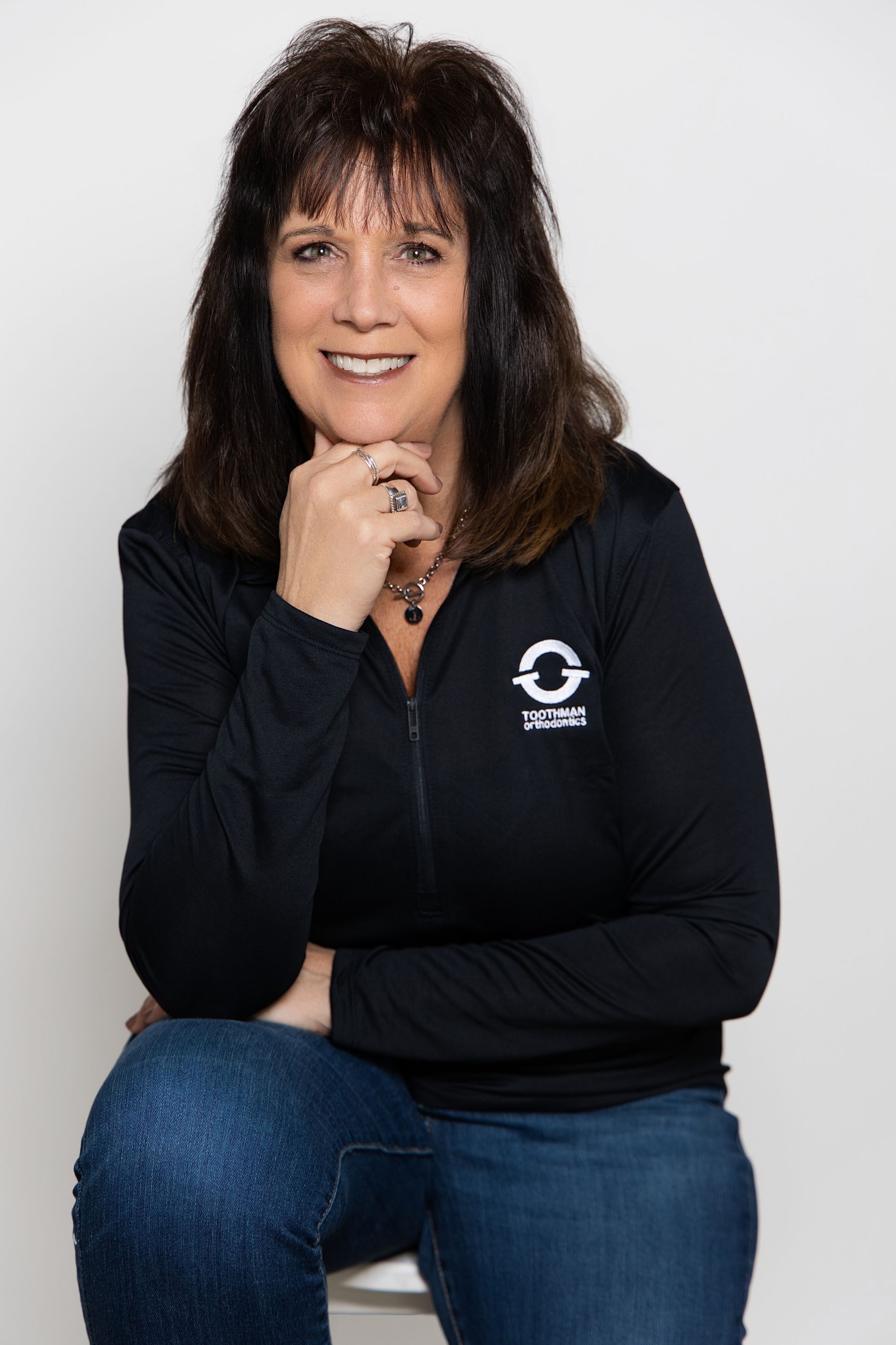 Woman with dark hair in a black top and jeans, seated with hand on chin against a light background