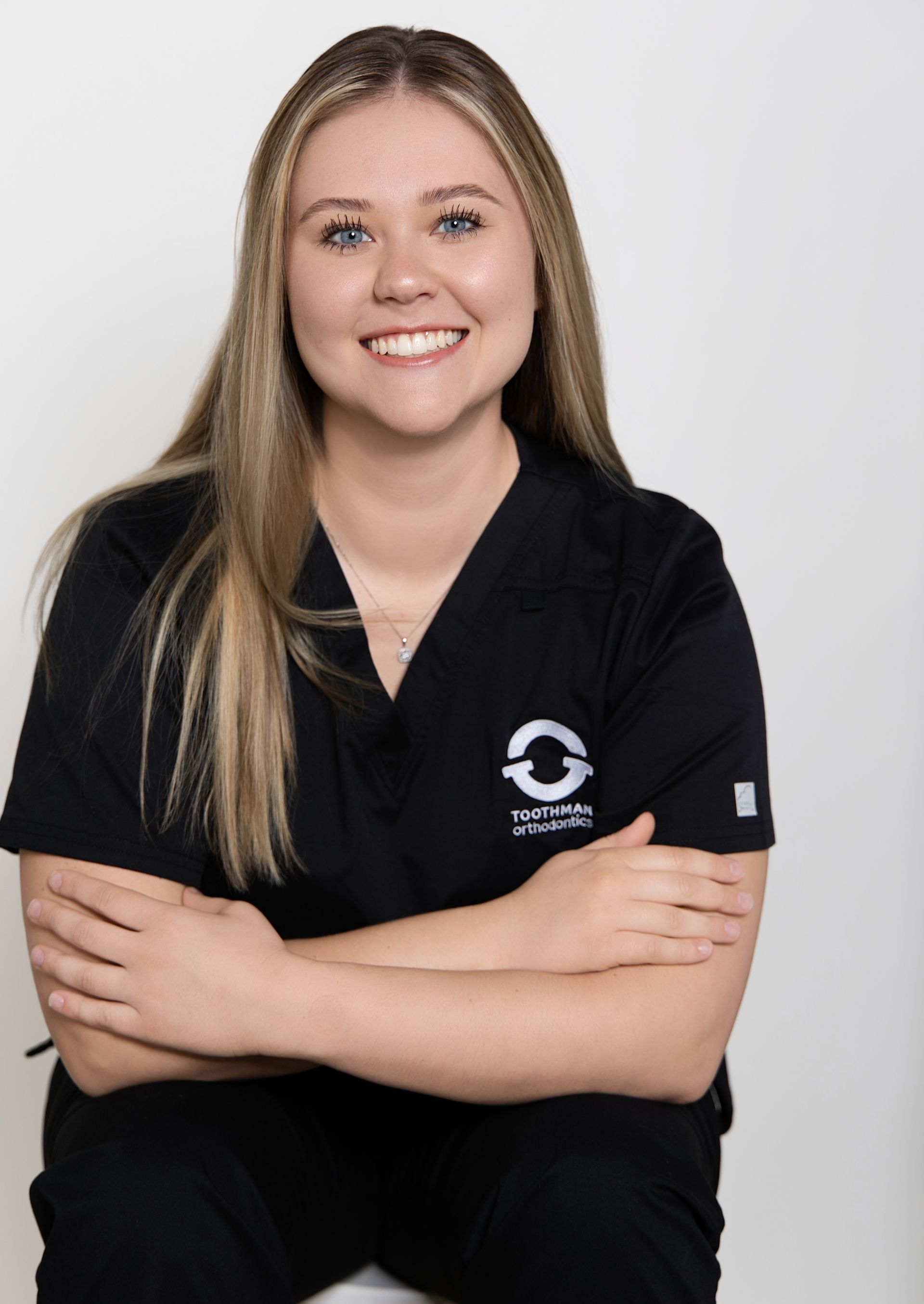 Smiling person in black scrubs, seated with arms crossed against a light gray background.