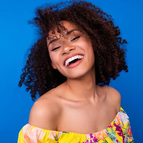 A woman with curly hair is smiling and wearing a yellow off the shoulder top.