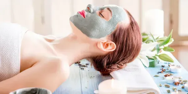Woman with a facial mask at a spa, resting on a pillow, with flowers and candles.