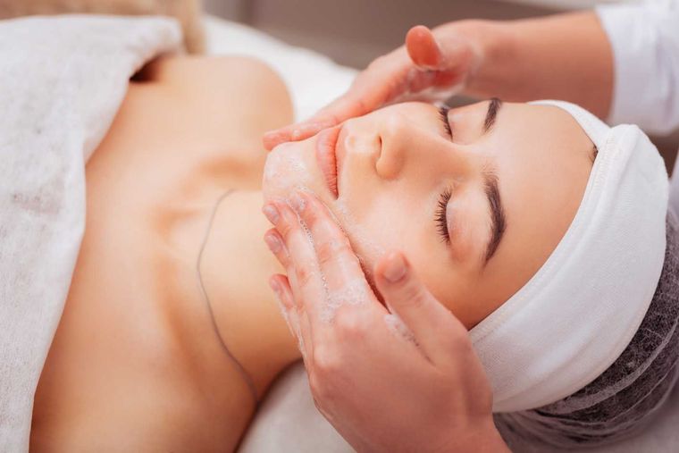 Woman receiving facial massage at a spa, eyes closed, hands on face, white towel and head wrap.