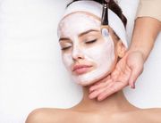 Woman with white facial mask, receiving treatment with brush, lying on a white surface.