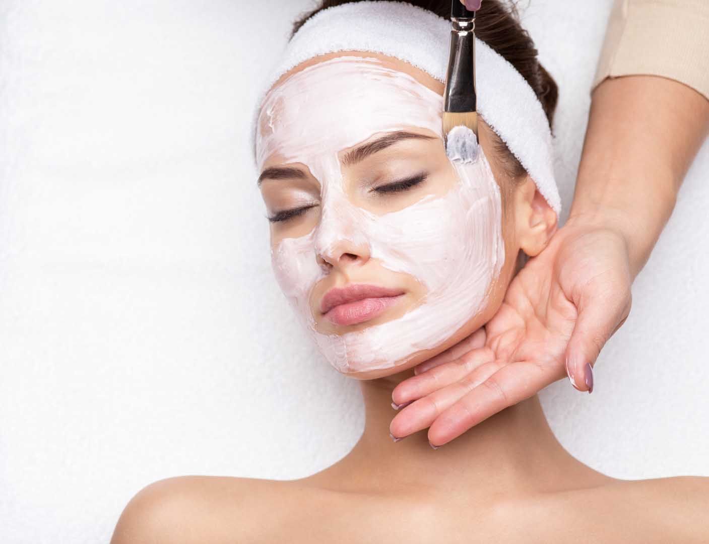 Woman with white facial mask, receiving treatment with brush, lying on a white surface.
