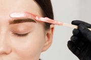 Eyebrow waxing in progress: esthetician applies pink wax above closed eye.