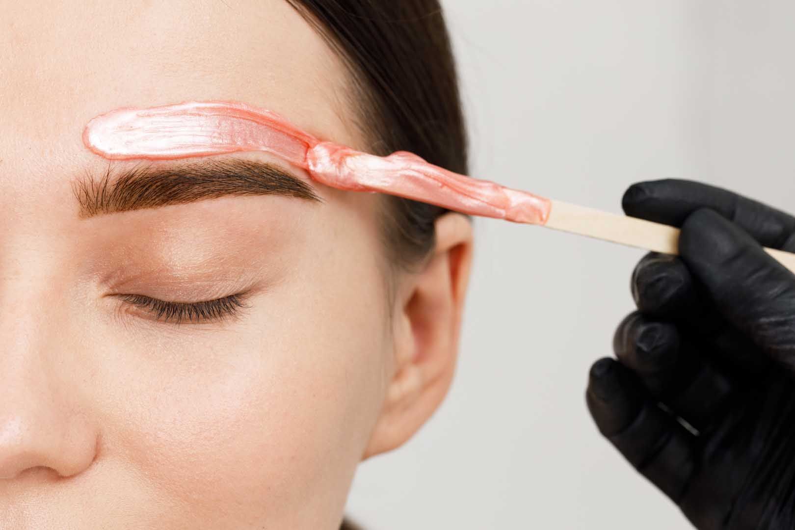 Eyebrow waxing in progress: esthetician applies pink wax above closed eye.
