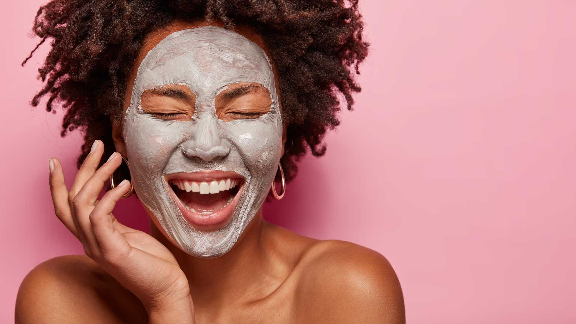 Woman with closed eyes, laughing, wearing a gray face mask against a pink background.