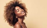 Smiling person with curly hair tilting their head against a beige background