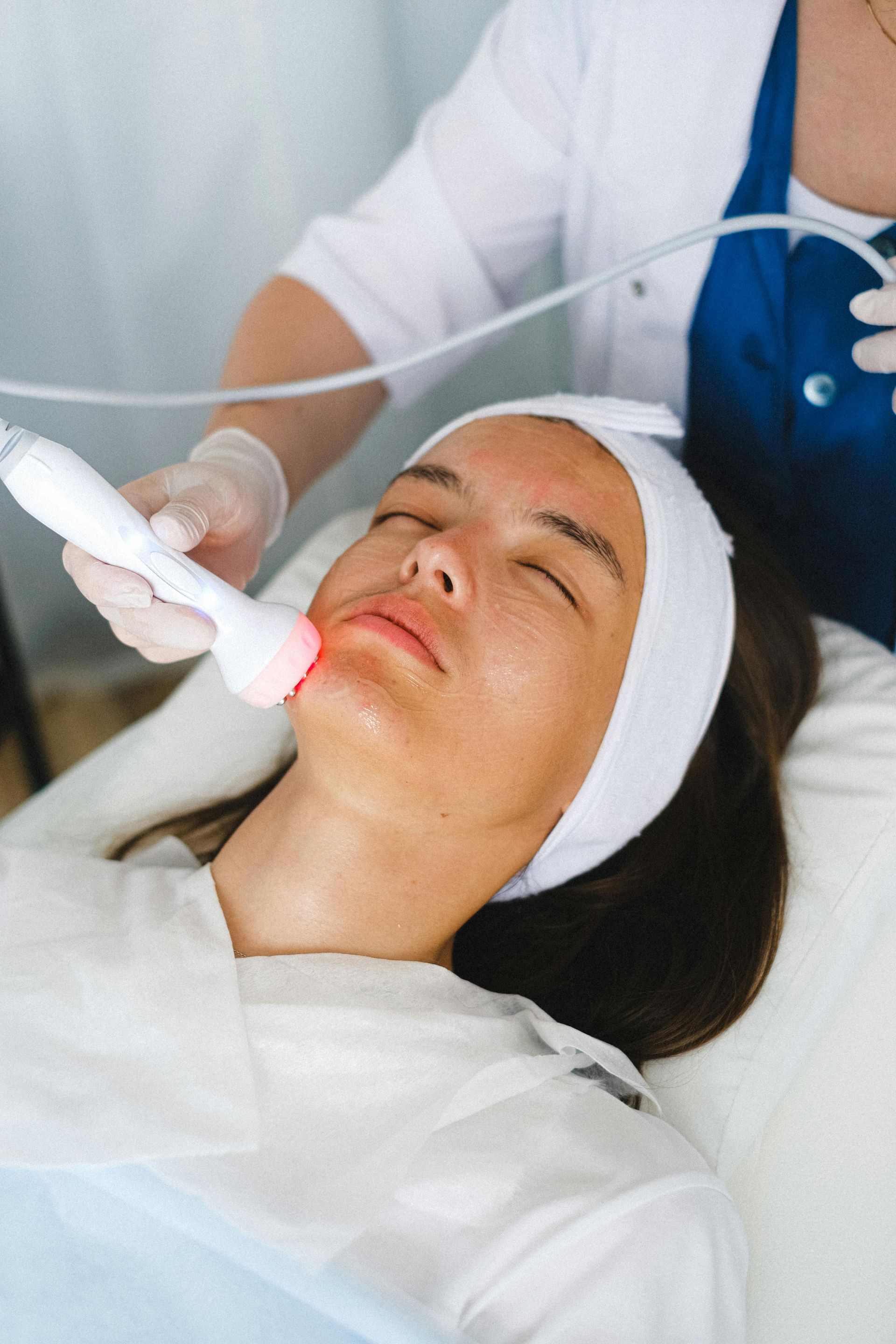 Woman receiving facial treatment; device near face, eyes closed, white headband.
