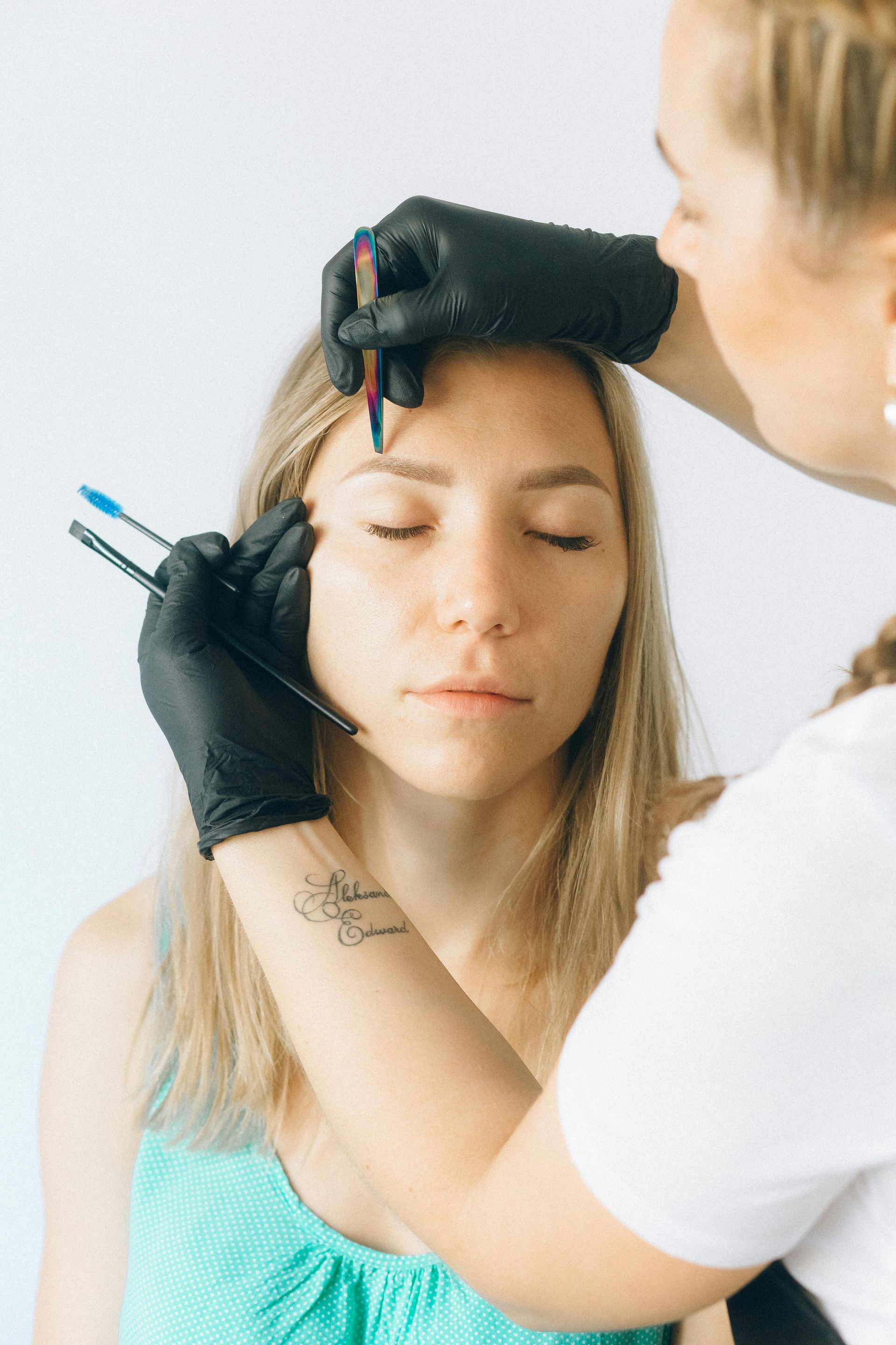 Eyebrow artist working on a woman's brow with tools, both wearing black gloves, in a bright setting.