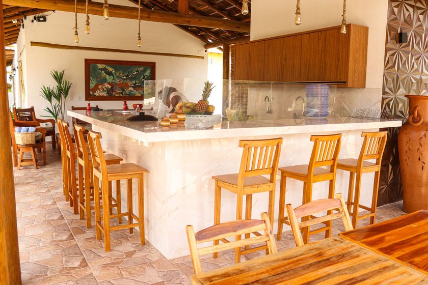 Cozinha tropical iluminada com ilha branca, banquetas de madeira e área de jantar aberta com teto de palha.