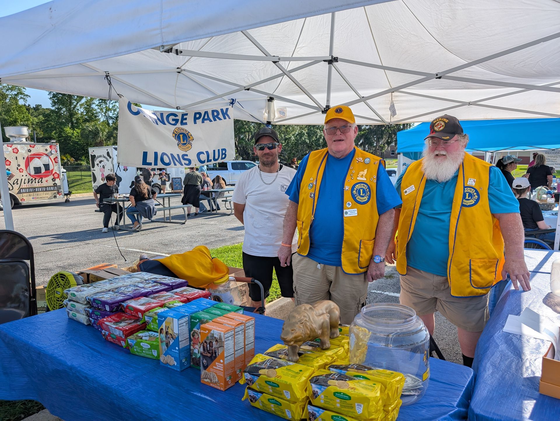 Three men at a Lions Club booth in a park, selling items under a white tent.