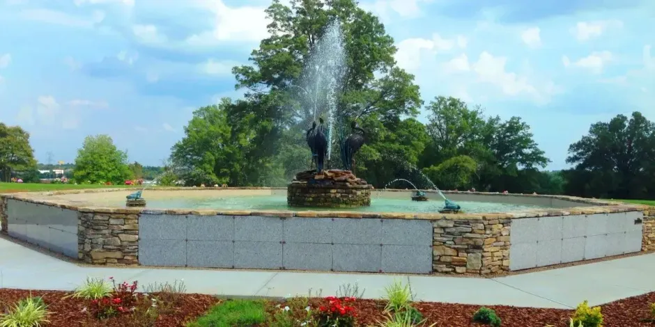 Public fountain spraying water in a stone plaza with trees and blue sky
