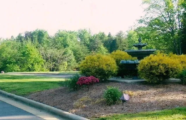 Landscaped garden with a stone fountain, pink flowers, shrubs, and trees beside a road