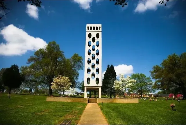 Tall white tower with circular openings in a green park under a blue sky with clouds