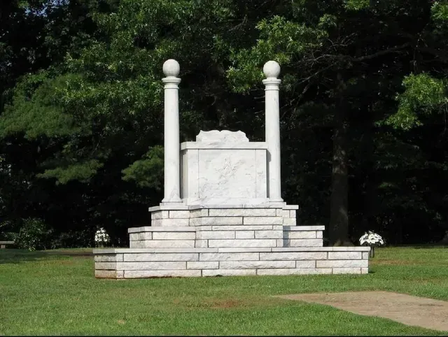 White stone monument with columns on a stepped base in a grassy park, backed by trees