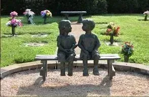 Two small bronze children statues sit on a bench in a grassy memorial garden.