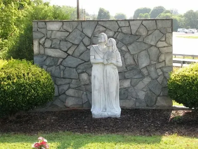 White statue in front of a stone wall, surrounded by green shrubs and grass.