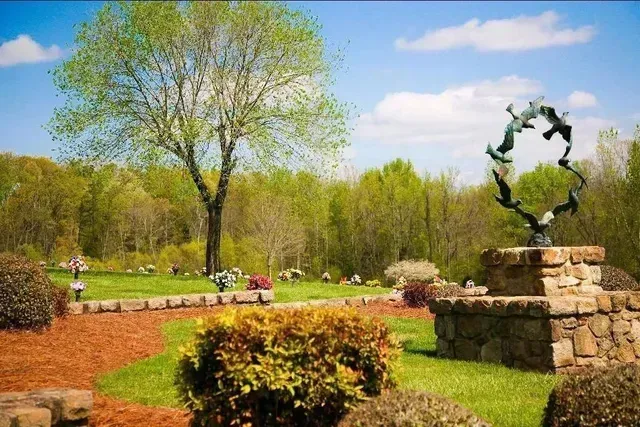 Sunny park with green trees, flower beds, a winding path, and a stone sculpture fountain.