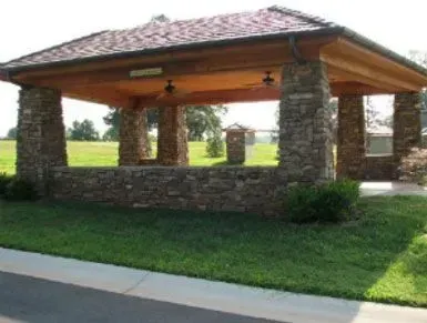 Stone pavilion with a wooden roof in a grassy park beside a sidewalk