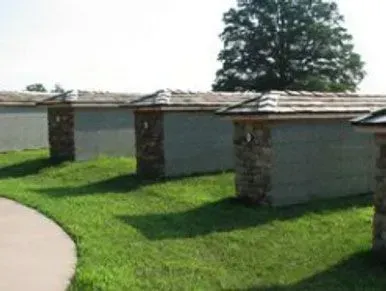 Row of small white-and-stone sheds beside a curved sidewalk and green lawn under a tree.