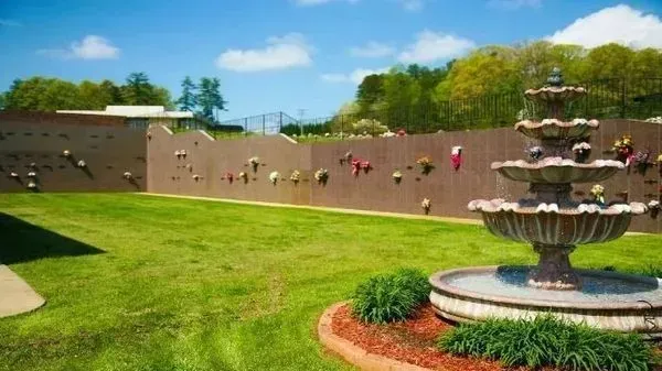 Sunny park with a tiered fountain, green lawn, tan wall, and colorful flowers under a blue sky