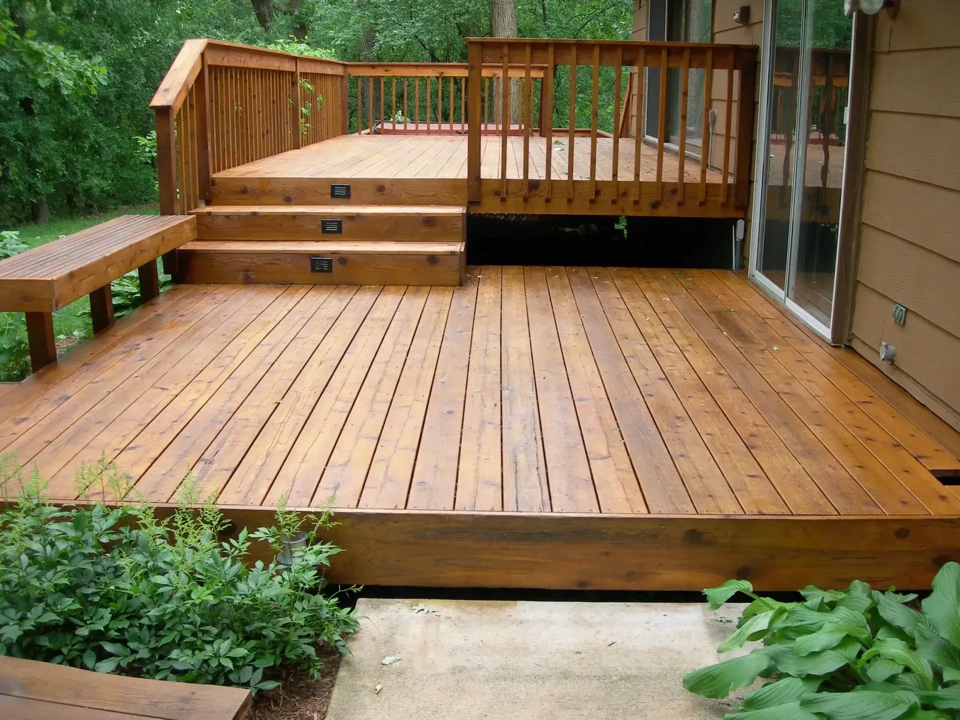 Wooden deck with stairs, bench, and railing; stained brown. Attached to a house with glass doors.