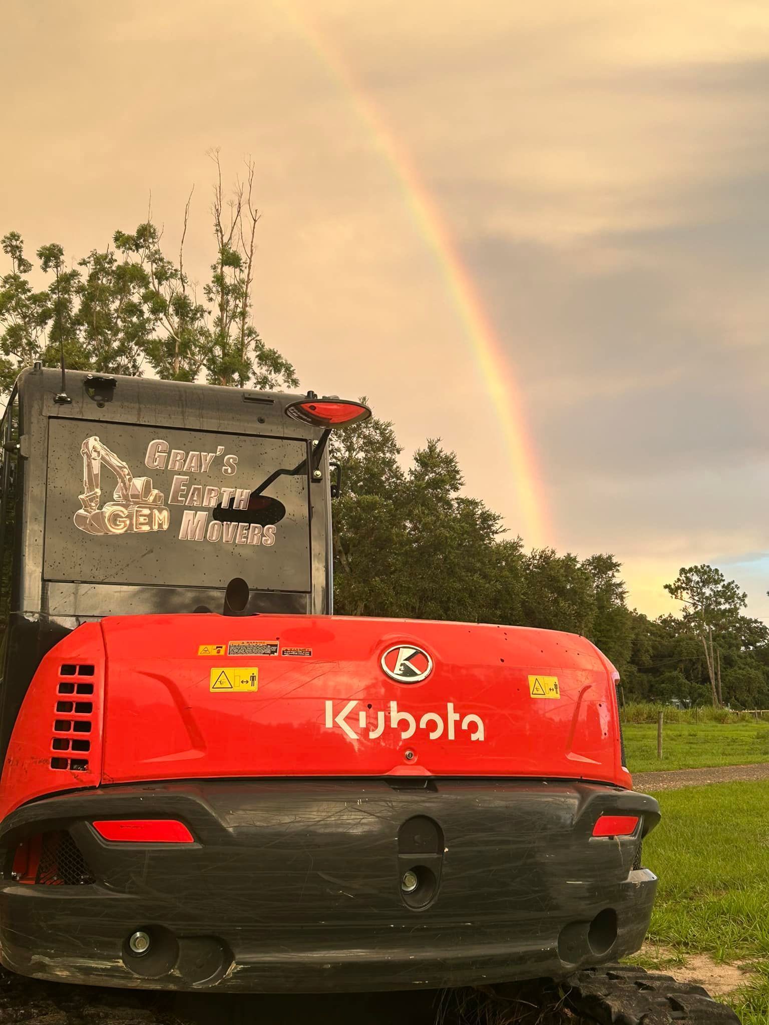 A red kubota excavator is parked in a field with a rainbow in the background.